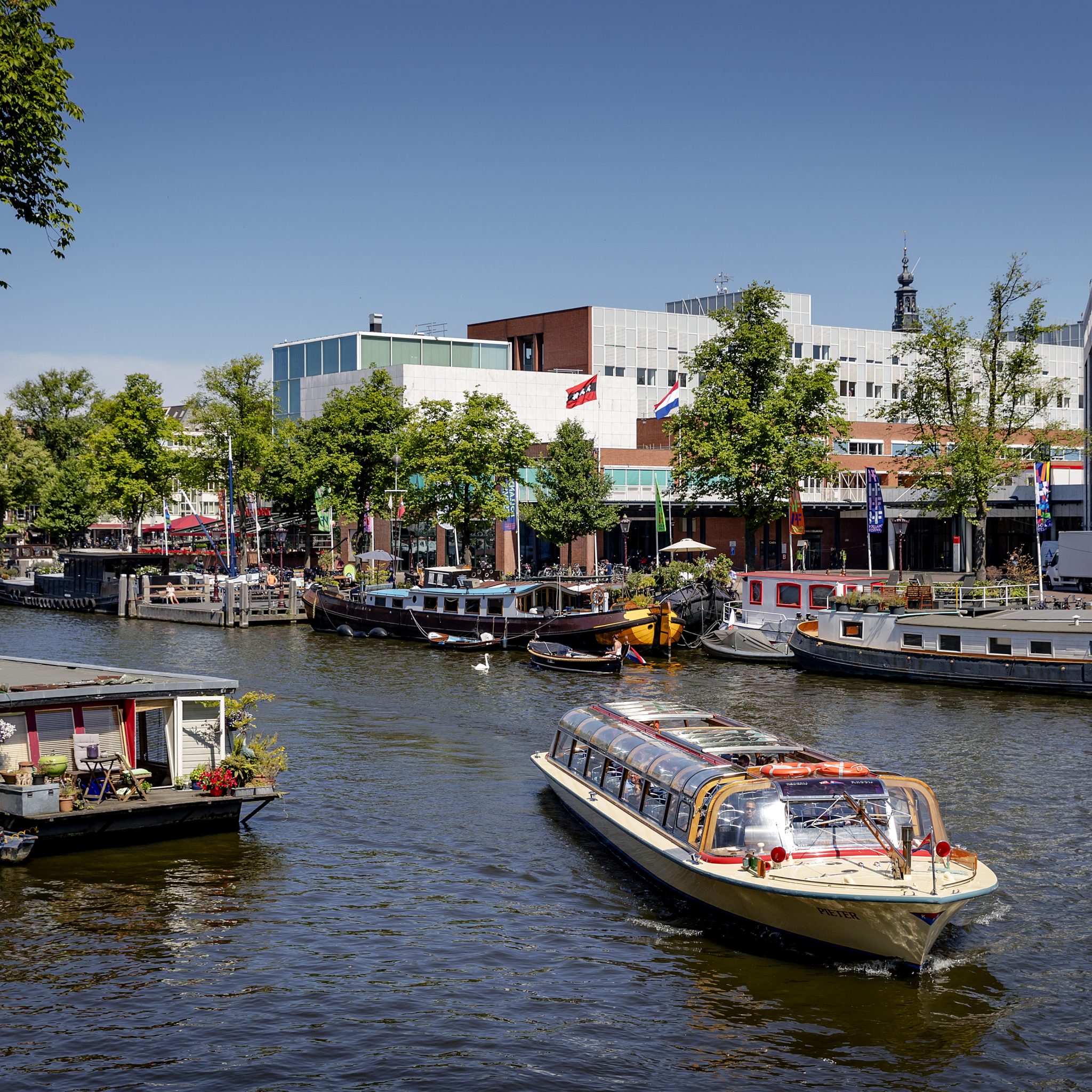 Een rondvaartboot vaart door de Amsterdamse grachten en passeert een woonboot. Op de achtergrond het Stadhuis in het centrum van Amsterdam.