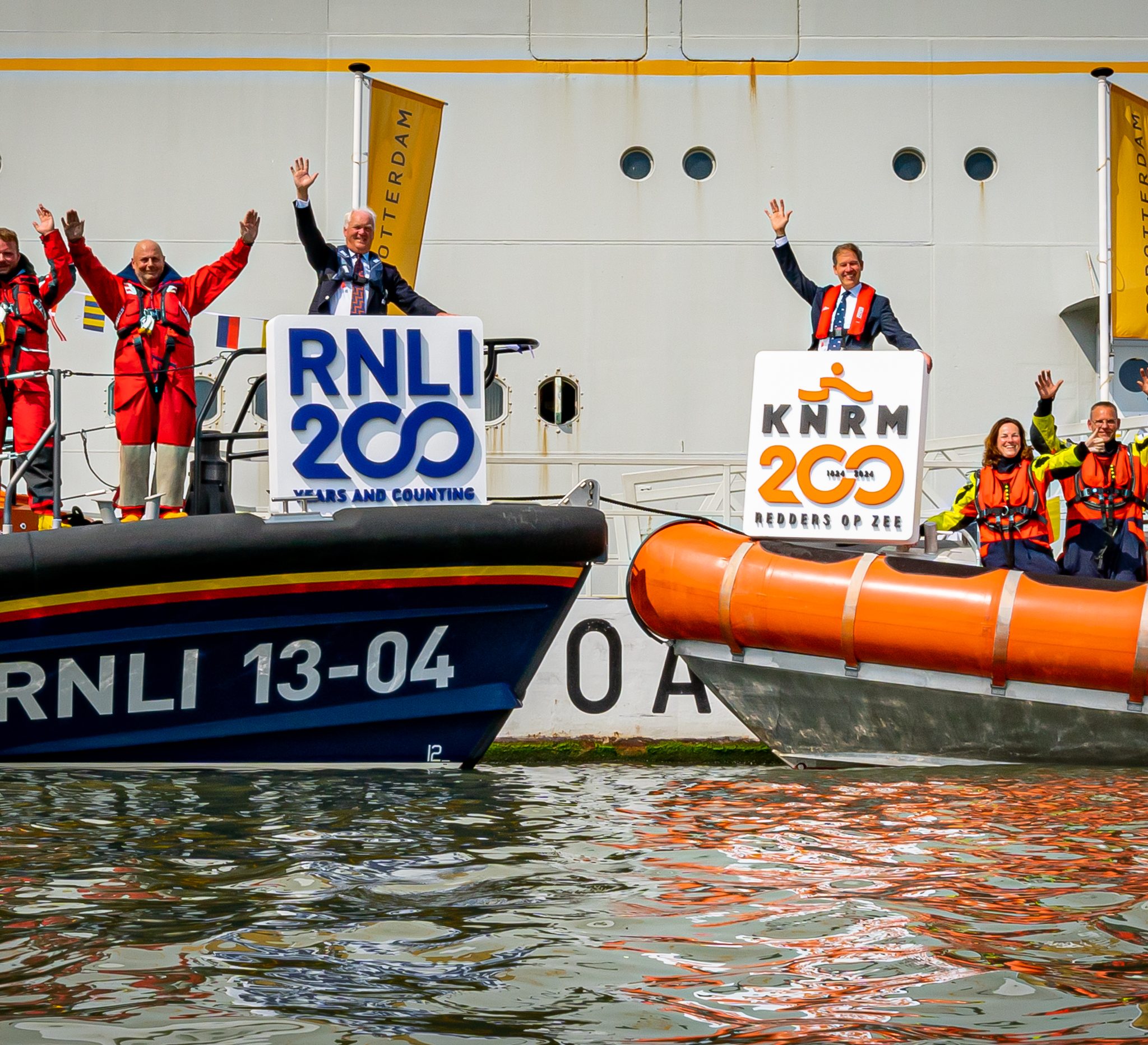 Directeur van de RNLI Mark Dowie (op de neus van de linker reddingboot) en Directeur Jacob Tas (op de neus van de rechter reddingboot) laten omringd door hun bemanning op het WMRC het gezamenlijke jubileumlogo zien.